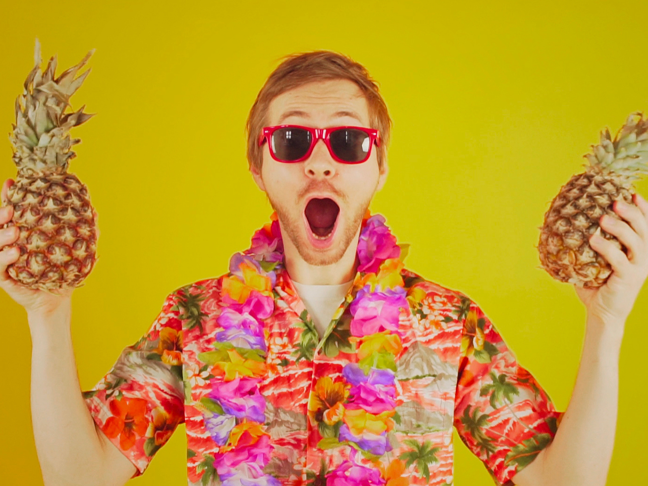 A man, who is wearing a pair of sunglasses, holding two pinapples with a surprised look
