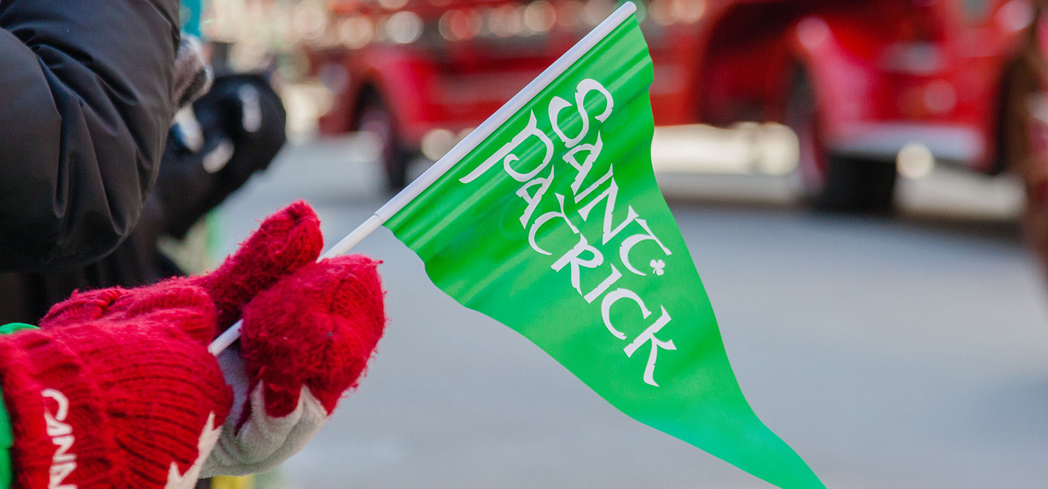 Canadian gloves holding a green Saint Patrick flag with a parade faded into the background.