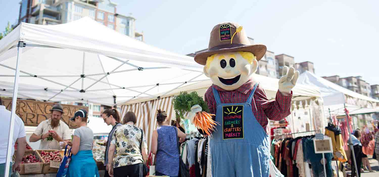 Farmers' Market mascot waving and holding carrots with people shopping in the background.